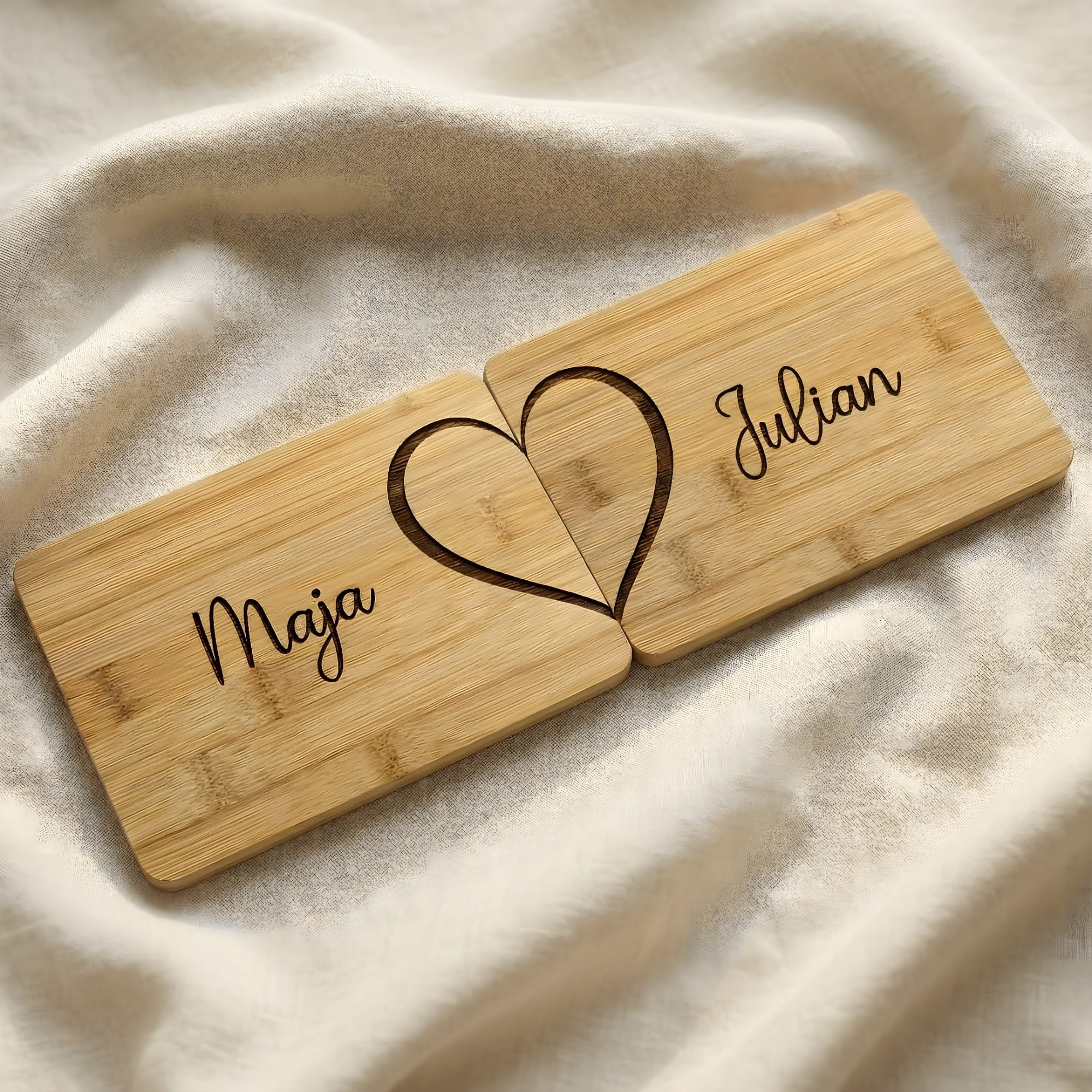 Zwei personalisierte Bambus-Frühstücksbrettchen als Paar-Set auf cremefarbenem Leinenstoff mit geteilter Herzgravur und Namen, Geschenk für Valentinstag oder Jahrestag.