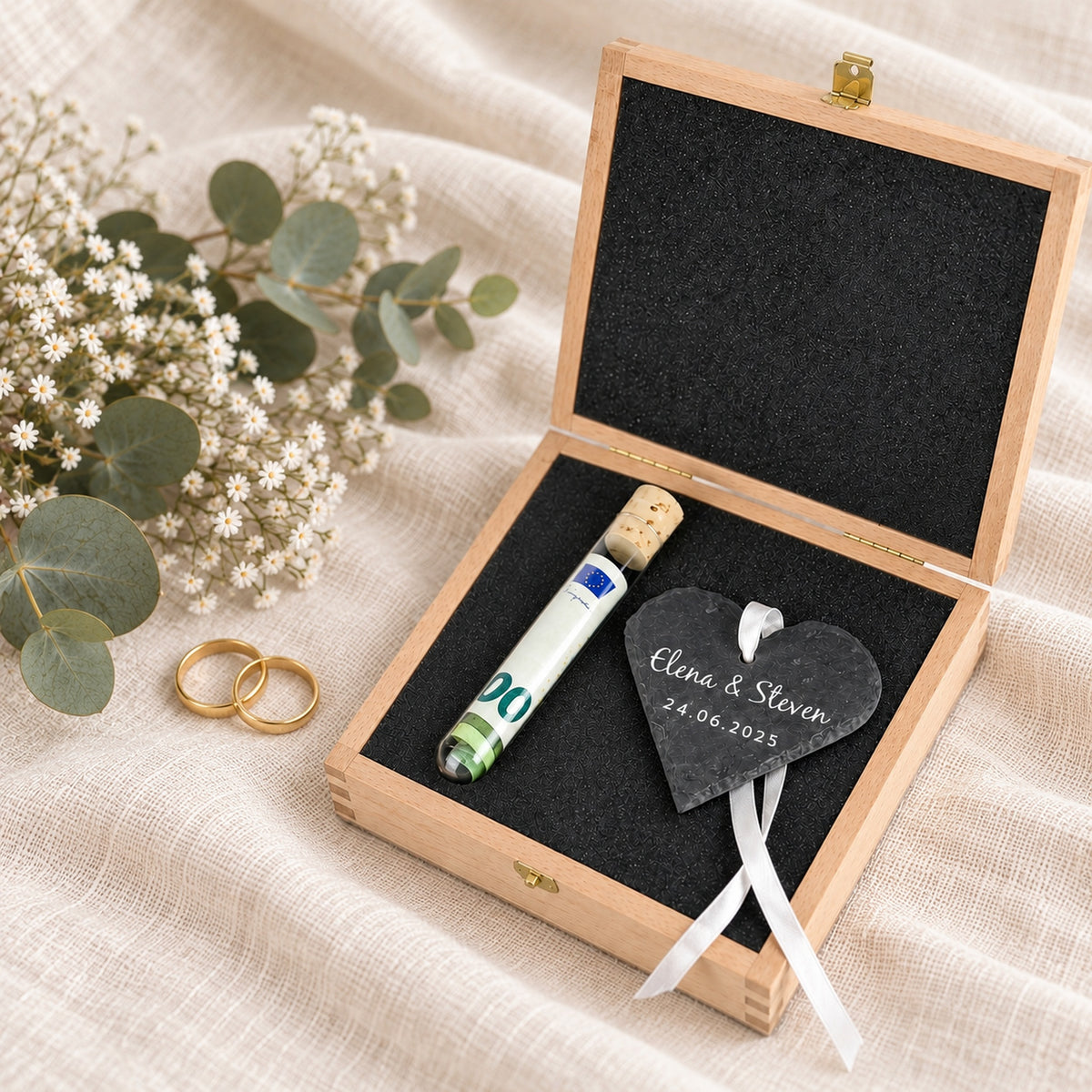 Geldgeschenk zur Hochzeit: offene Holzbox mit Reagenzglas für Geldscheine und graviertem Schieferherz, daneben zwei Eheringe und Eukalyptus