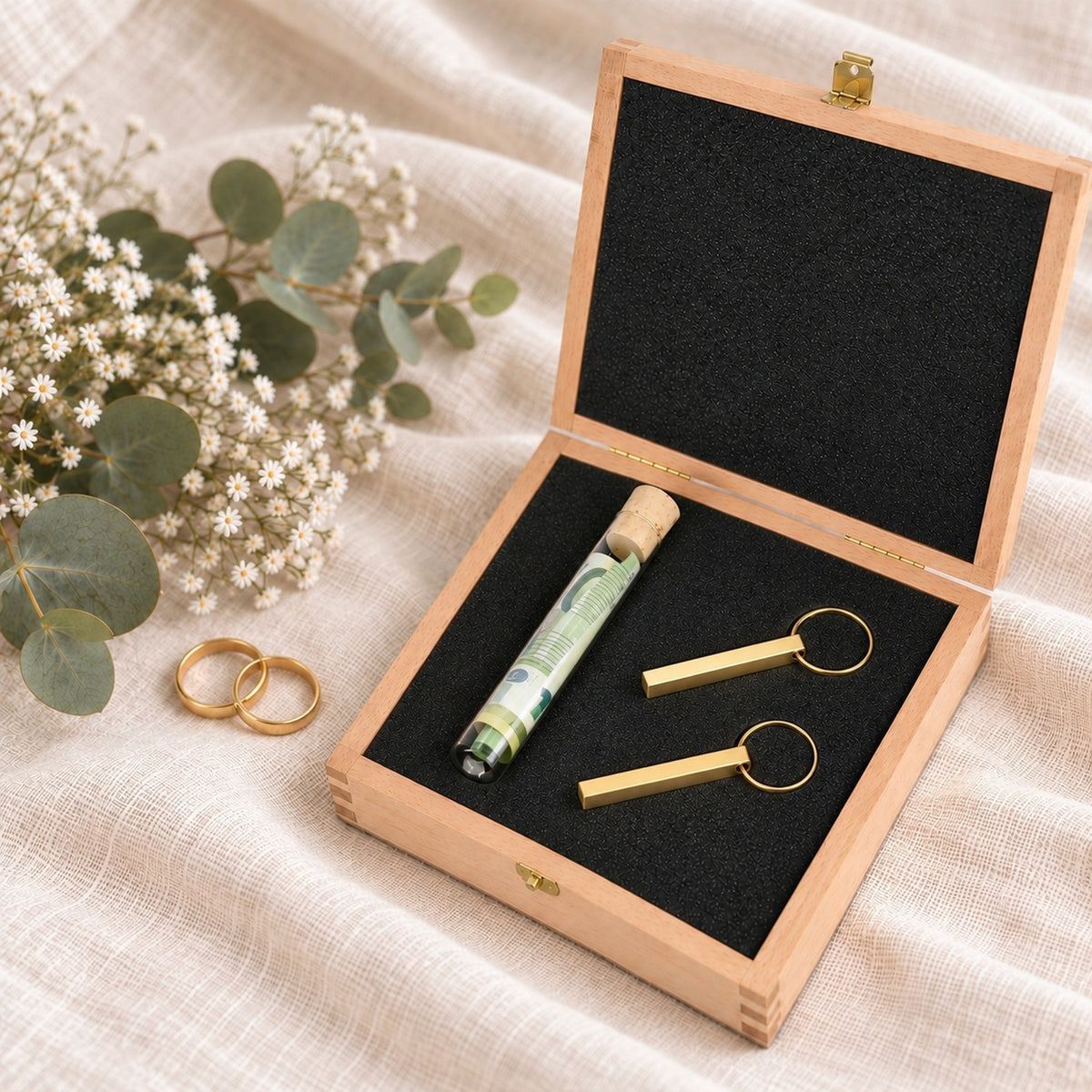 Geldgeschenk zur Hochzeit: offene Holzbox mit Reagenzglas für Geldscheine und zwei goldfarbenen Schlüsselanhängern, daneben zwei Eheringe und Eukalyptus